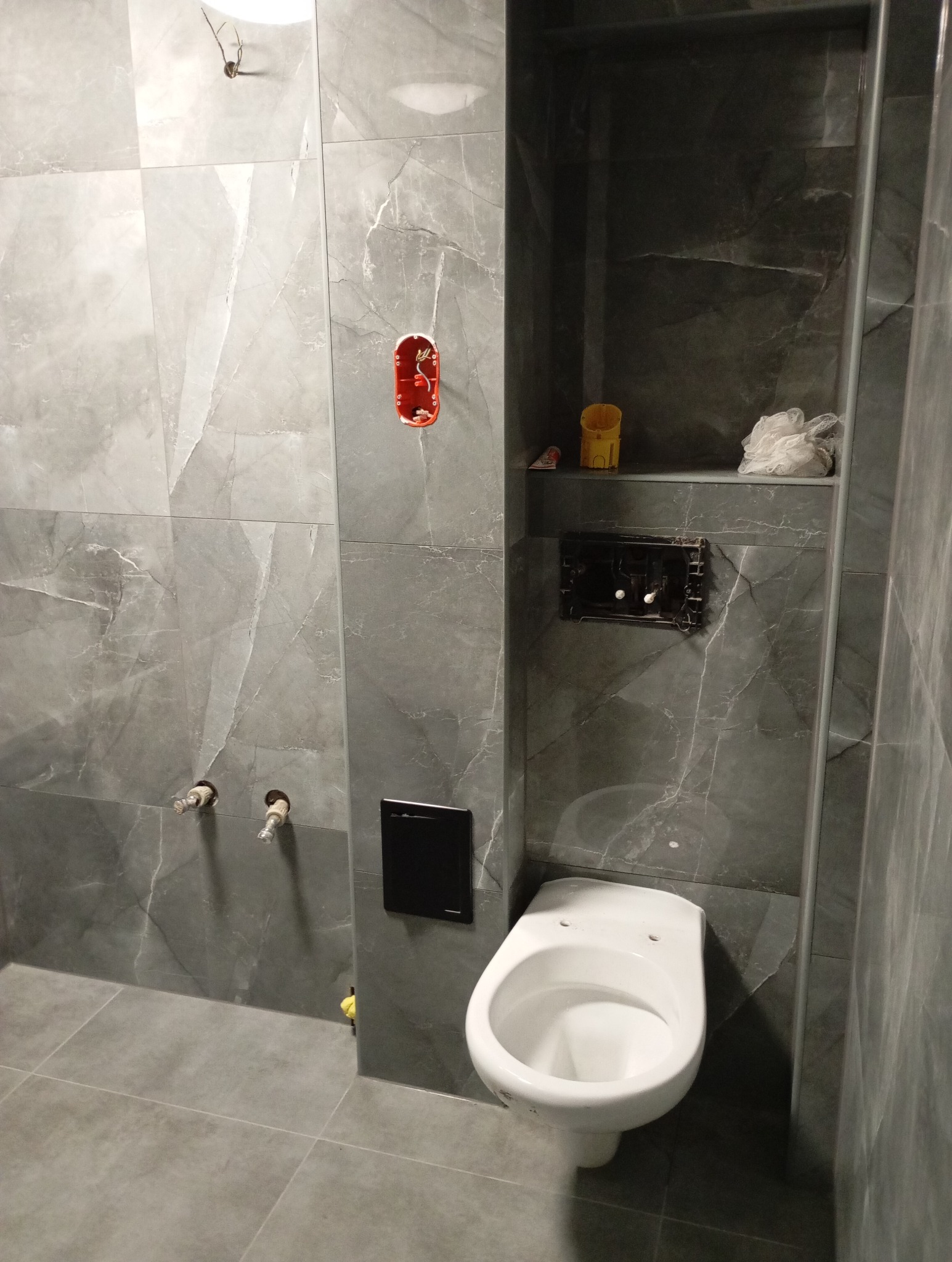 Niedokończona łazienka z szarymi płytkami imitującymi marmur, wisząca toaleta bez deski, puszka elektryczna wystająca ze ściany i niezamaskowane rury doprowadzające wodę.