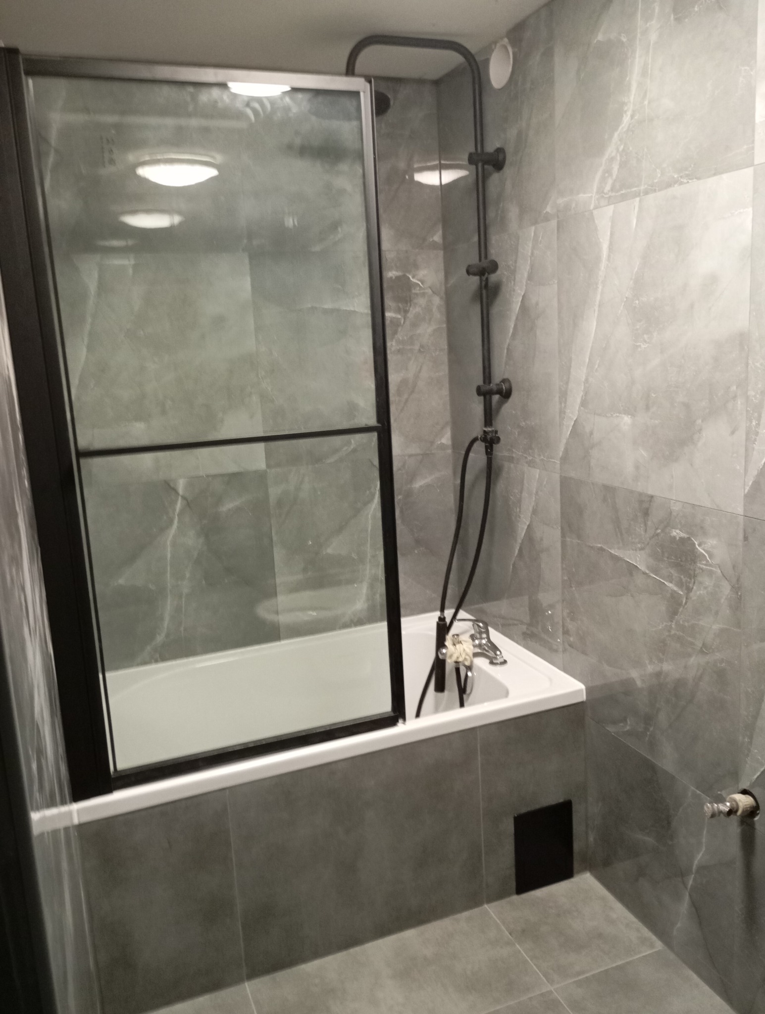 Szara łazienka z wanną obudowaną płytkami, czarną kabiną prysznicową i baterią natryskową w stylu retro.