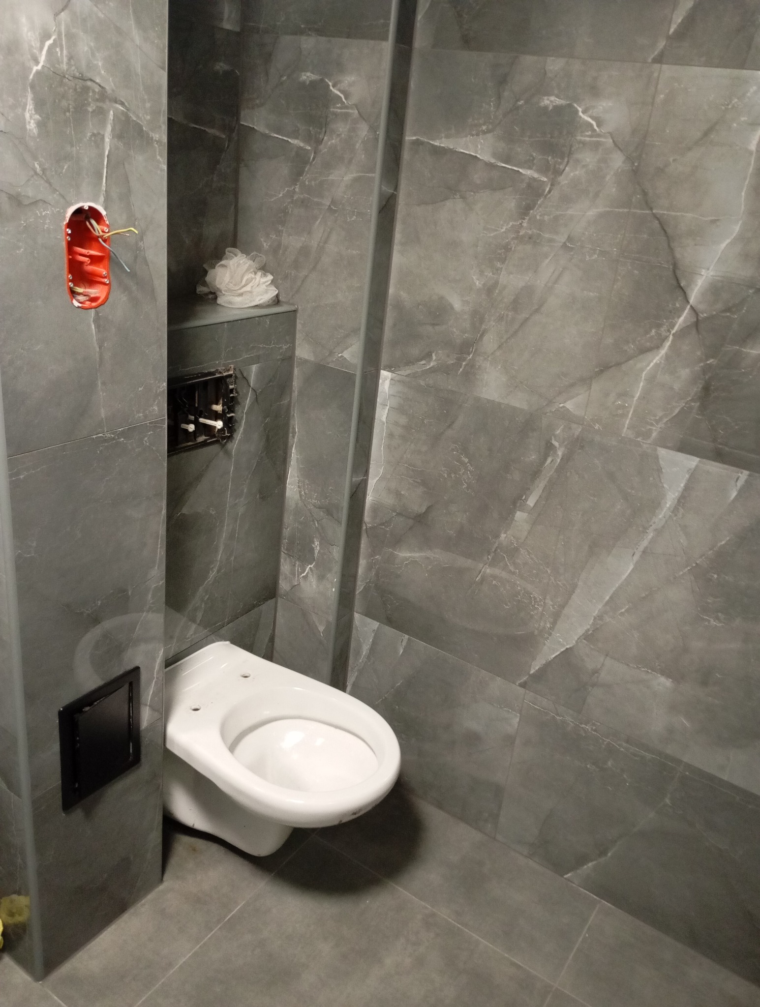 Szara łazienka z marmurowymi płytkami, częściowo wykończona, z widoczną instalacją elektryczną i stelażem podtynkowym do WC.