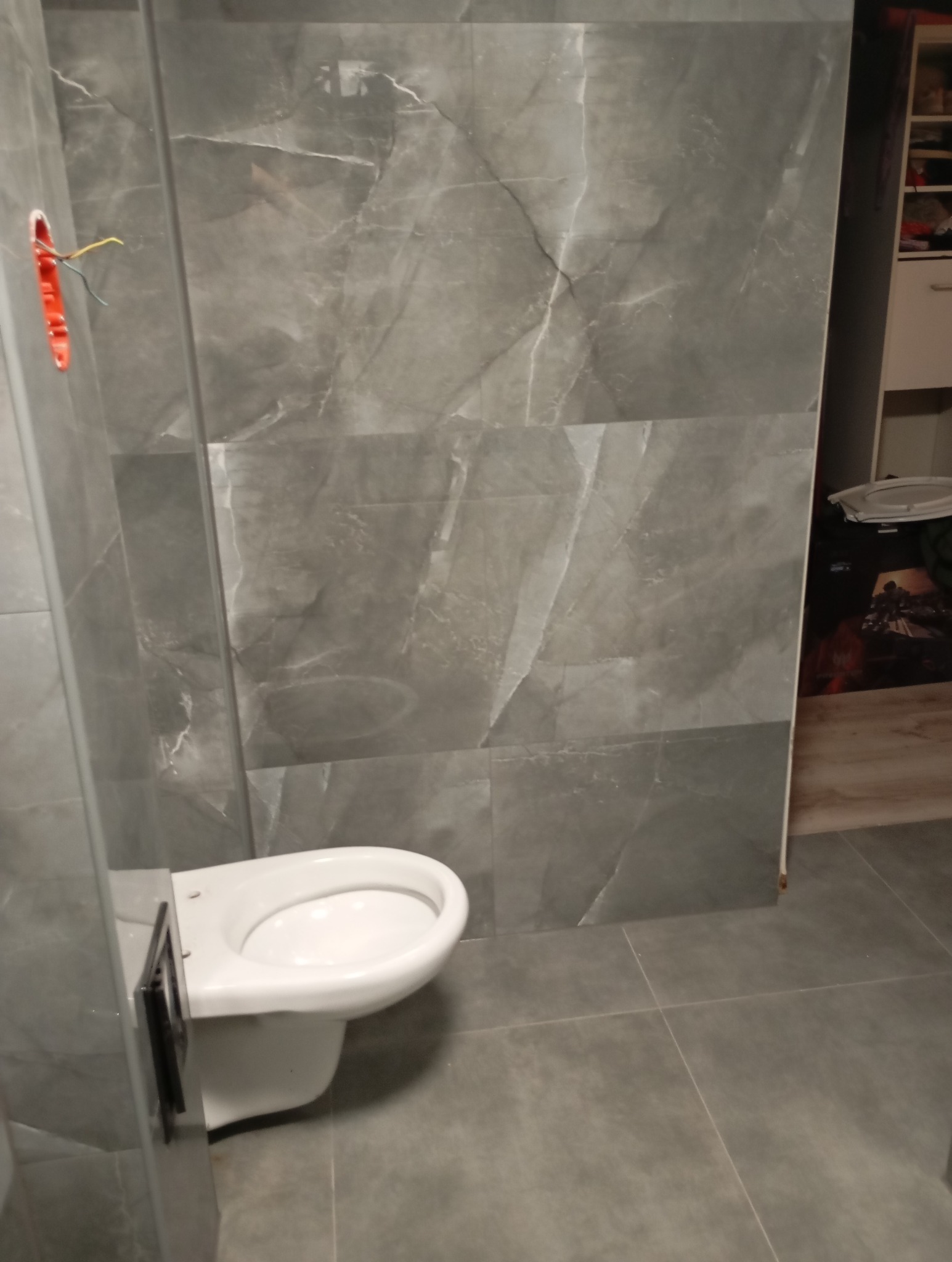 Świeżo wyremontowana łazienka z szarymi płytkami imitującymi marmur, podwieszana toaleta bez deski, widoczne kable wystające ze ściany.