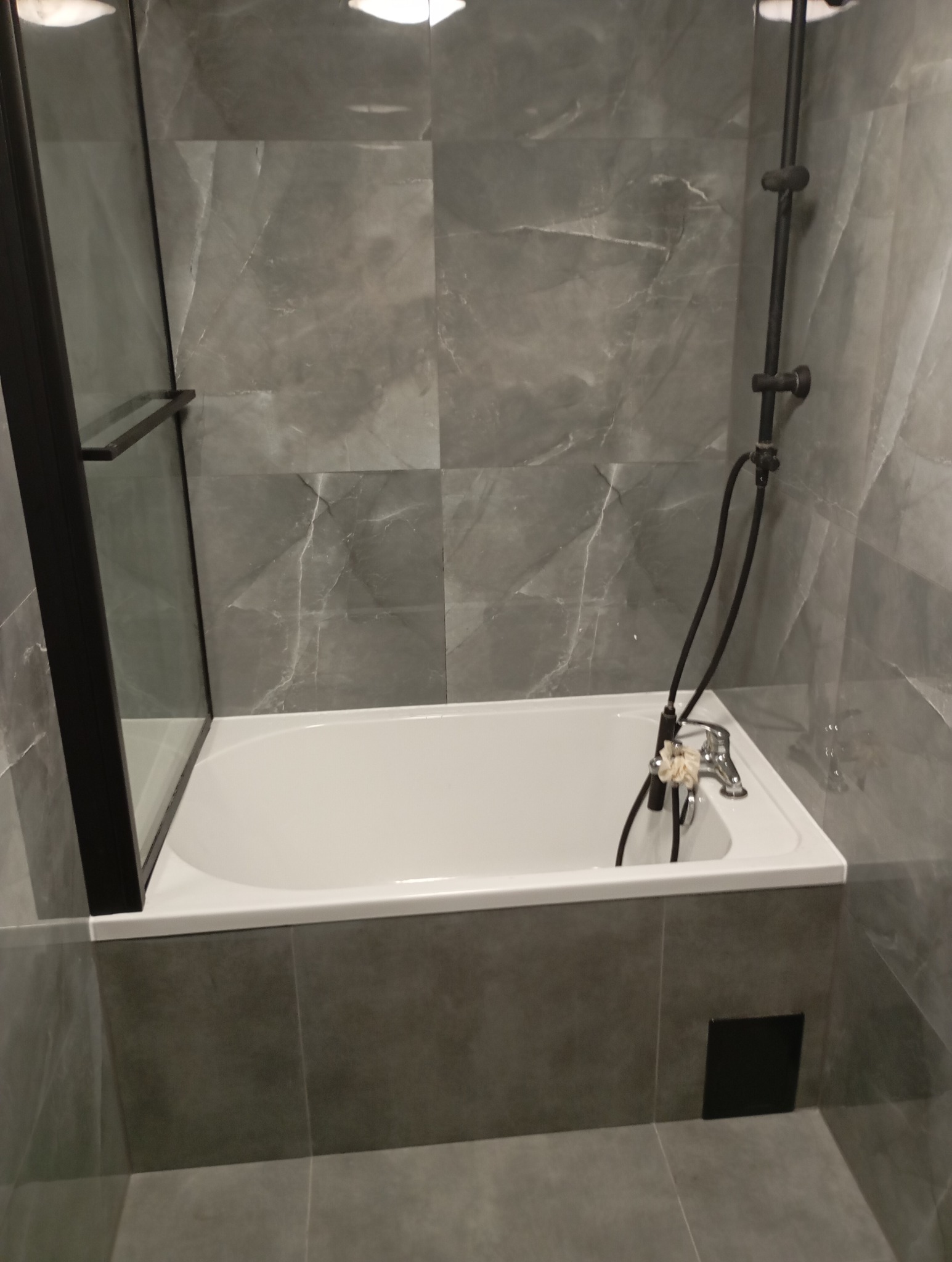 Wykończona łazienka z wanną z czarnym parawanem nawannowym, szarą armaturą i płytkami imitującymi marmur.