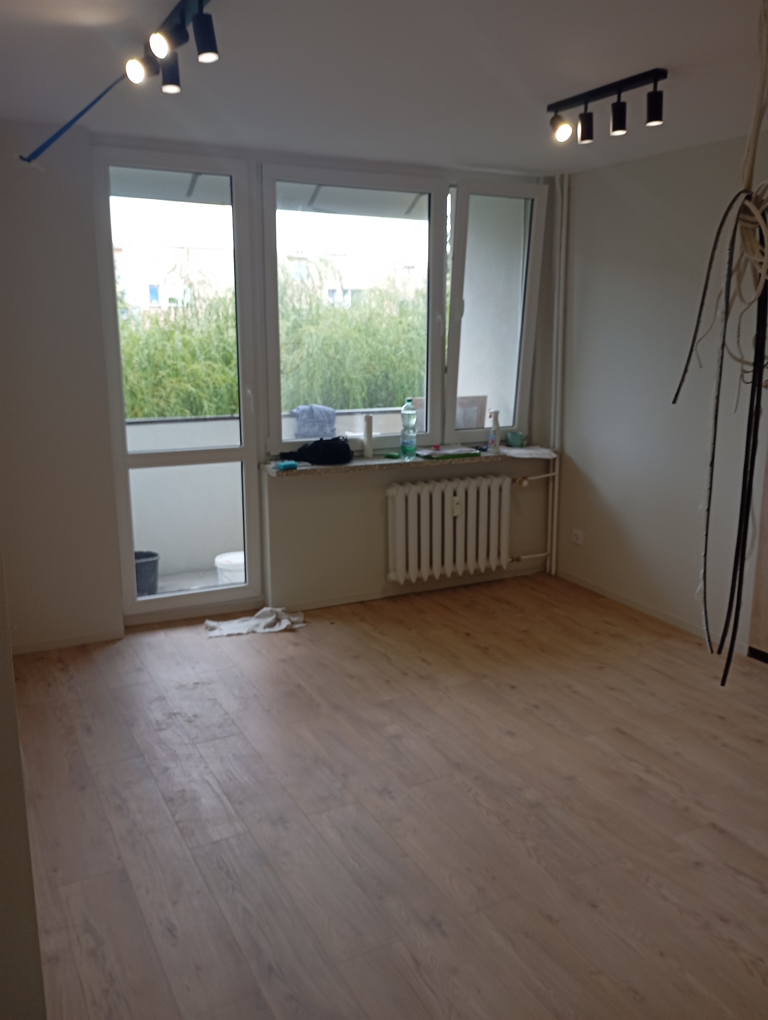 Pusty pokój po położeniu paneli podłogowych, widoczne okno z widokiem na zieleń, grzejnik pod oknem i lampy sufitowe.