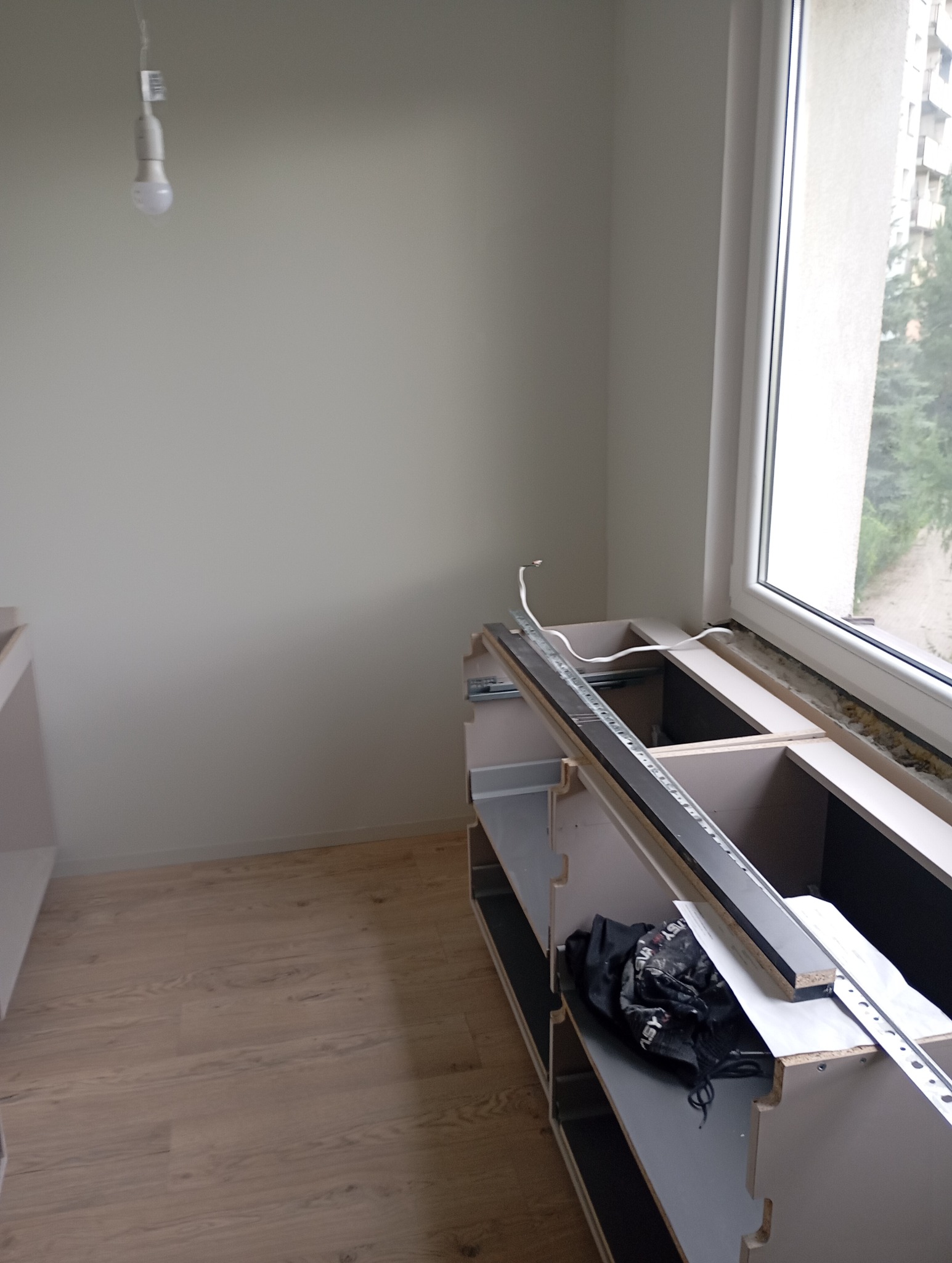 Montaż szafek kuchennych pod oknem, widoczne szyny montażowe i przewody elektryczne, surowy stan wykończenia.