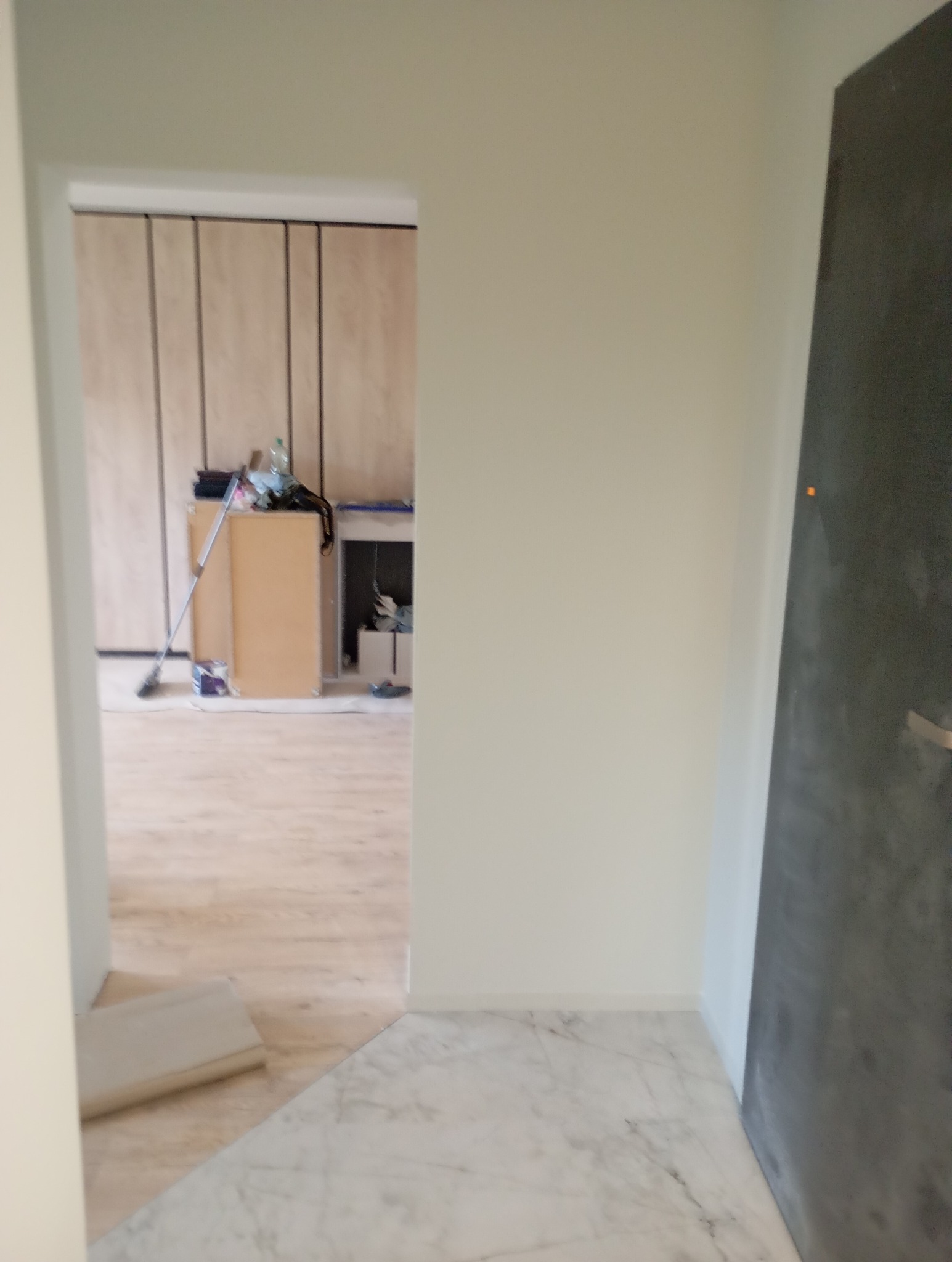 Widok przez drzwi na remontowane pomieszczenie z drewnianą podłogą i marmurową posadzką przy wejściu, widoczne narzędzia i materiały budowlane.