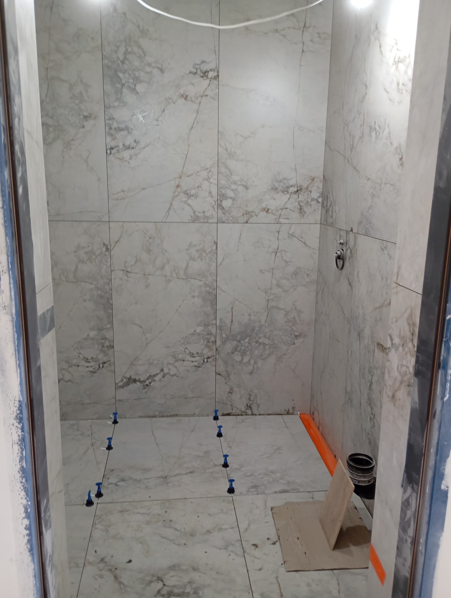 Układanie płytek imitujących marmur w łazience, widoczne systemy poziomowania płytek na podłodze, pomarańczowe zabezpieczenia krawędzi, surowe wykończenie.