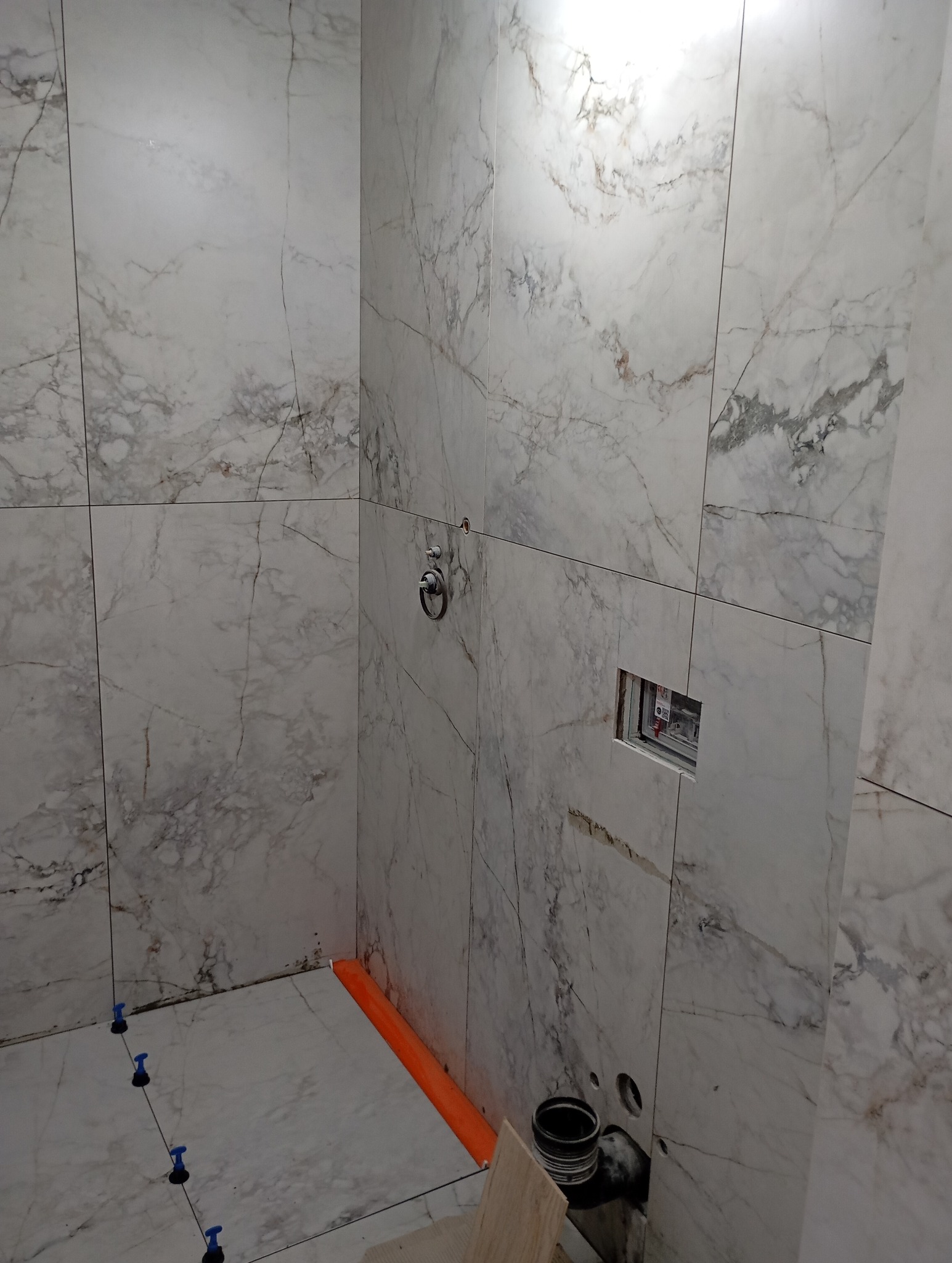 Łazienka w trakcie kładzenia płytek marmurkowych, widoczne krzyżyki poziomujące, rura odpływowa oraz otwór na instalację elektryczną.