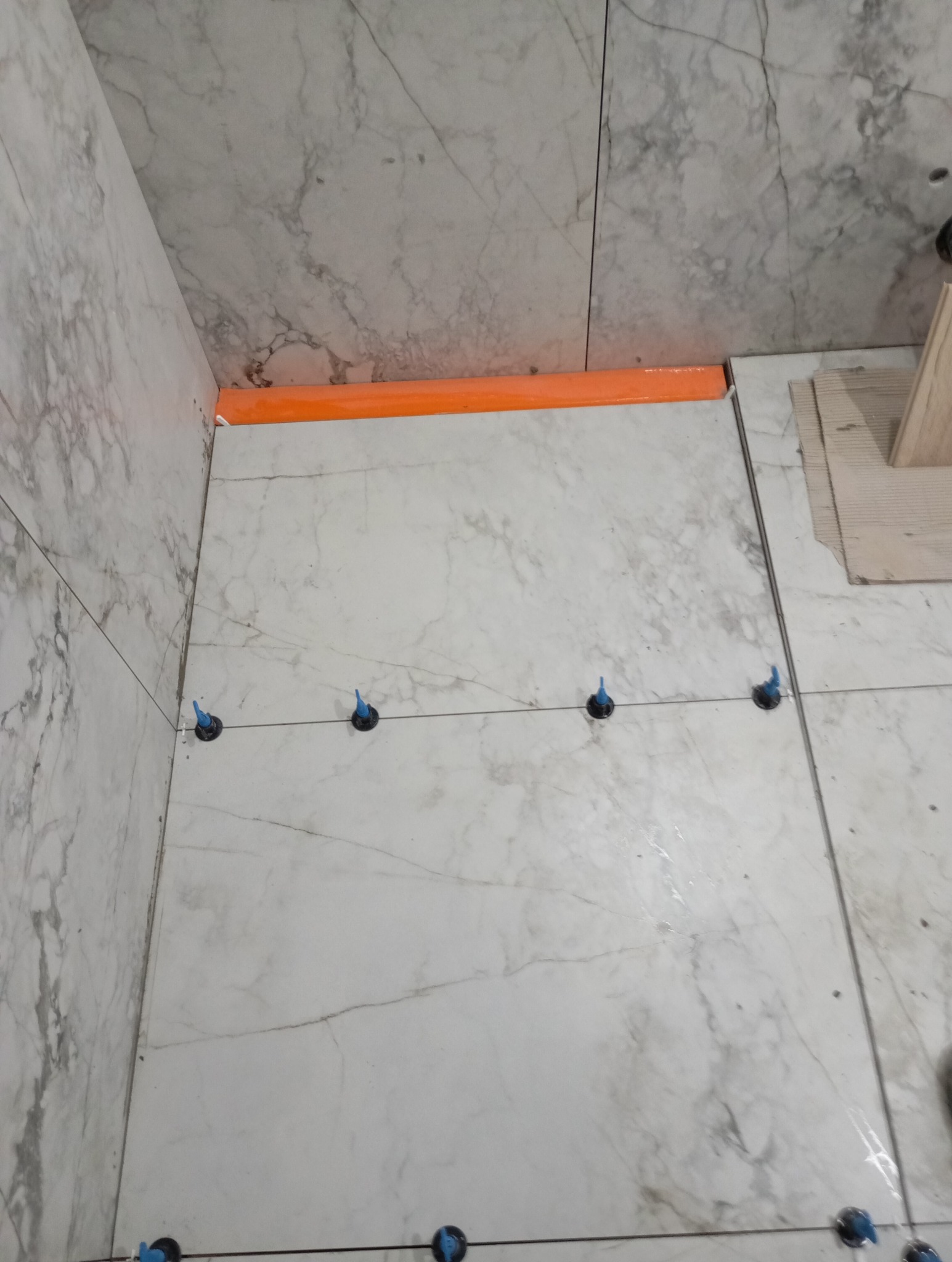 Układanie płytek imitujących marmur na podłodze w rogu pomieszczenia, widoczne niebieskie kliny do poziomowania i pomarańczowa listwa wykończeniowa.