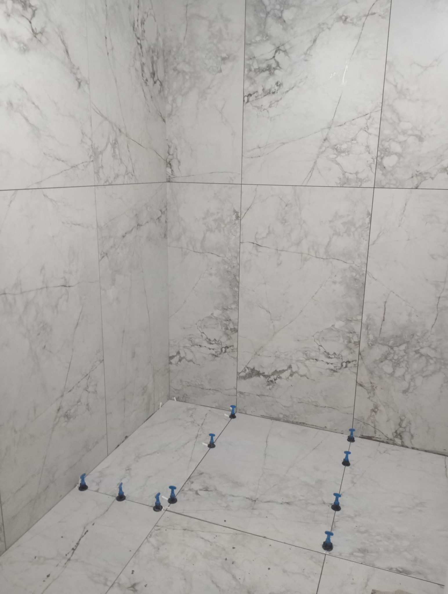 Białe płytki imitujące marmur ułożone w rogu łazienki, z niebieskimi klipsami poziomującymi, widoczne fugi.