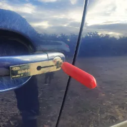 Otwieranie zamka w niebieskim samochodzie za pomocą specjalistycznego wytrychu z czerwonym uchwytem, marki COSO HU66, z widocznym odbiciem chmur na karoserii.