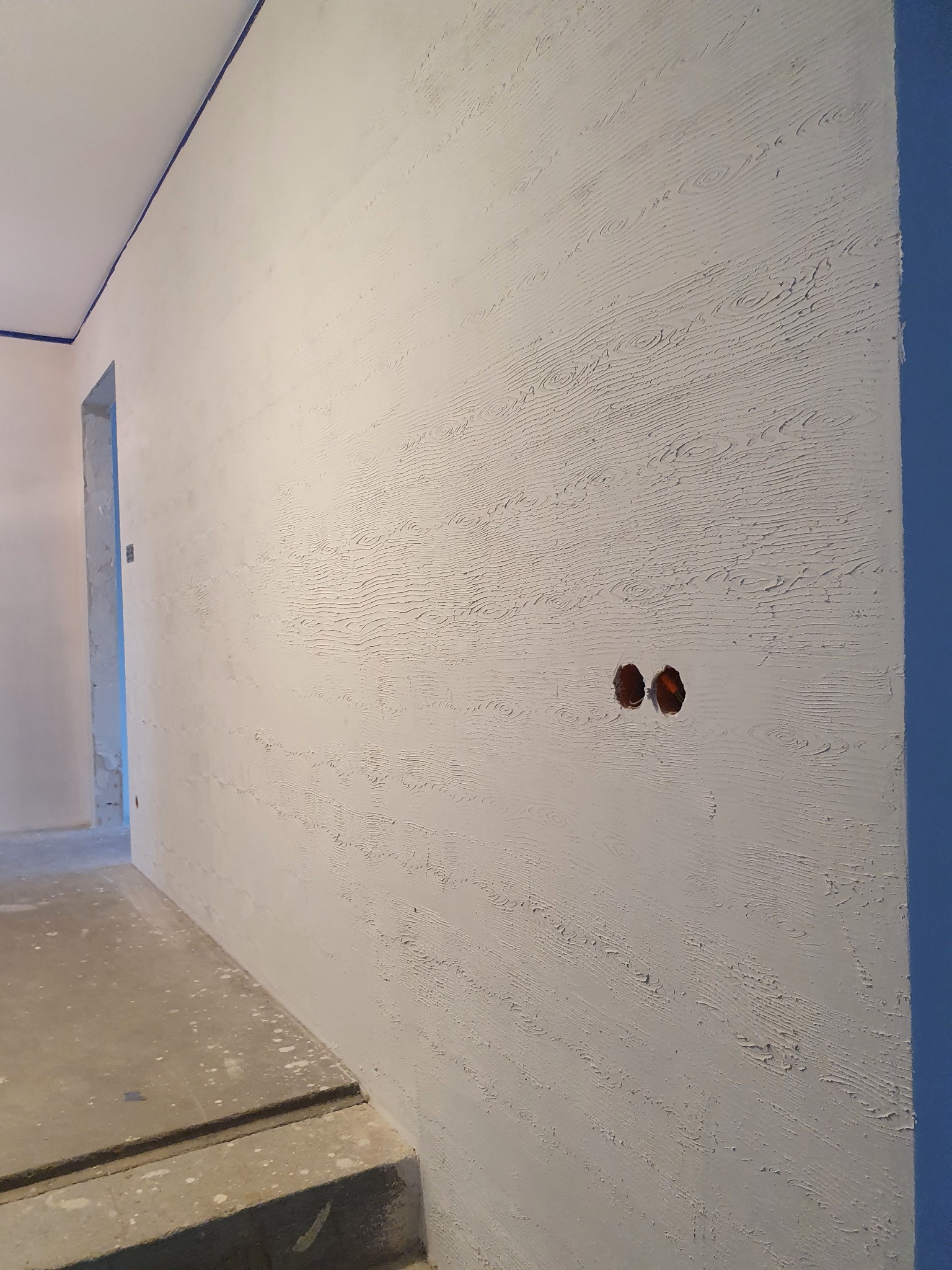 Ściana z fakturą imitującą drewno po nałożeniu gładzi, widoczne otwory na gniazdka elektryczne, zabezpieczona taśmą malarską listwa sufitowa.