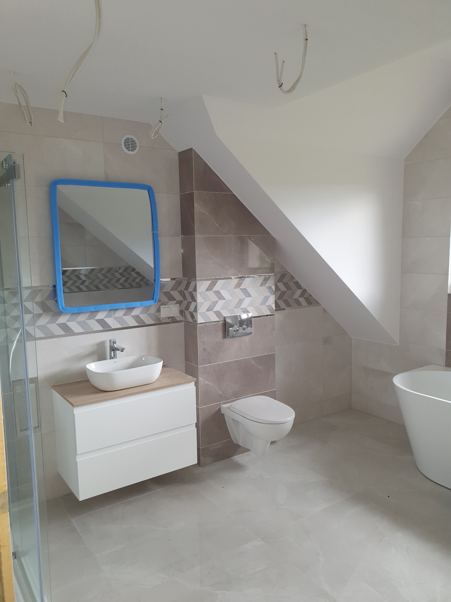 Nowoczesna łazienka na poddaszu z umywalką nablatową, lustrem w niebieskiej ramie zabezpieczonej folią, wiszącą toaletą i fragmentem wanny, wykończona płytkami w odcieniach beżu i brązu...