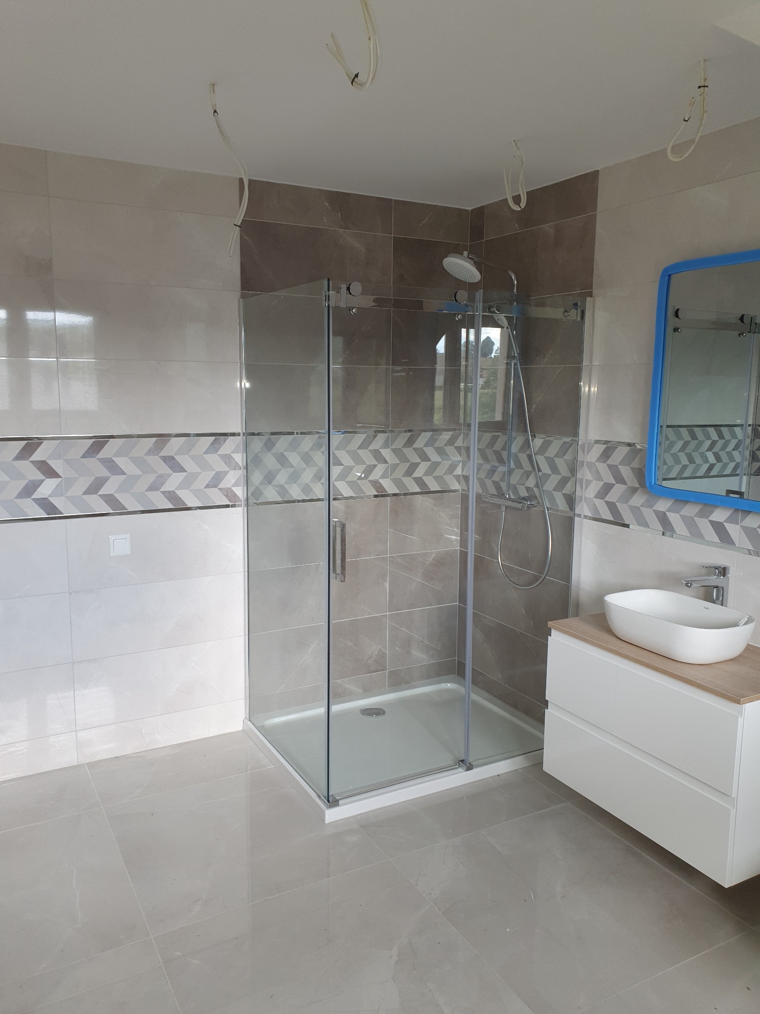 Nowoczesna łazienka z przeszkloną kabiną prysznicową, umywalką nablatową na białej szafce i lustrem w niebieskiej ramie, wykończona płytkami w odcieniach beżu i brązu z dekoracyjnym pasem, widoczne...