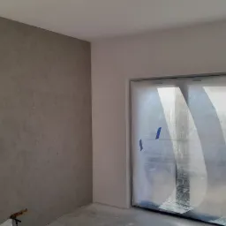 Ściana z efektem dekoracyjnym w odcieniach szarości, zabezpieczone okno folią malarską, widoczna drabina malarska.