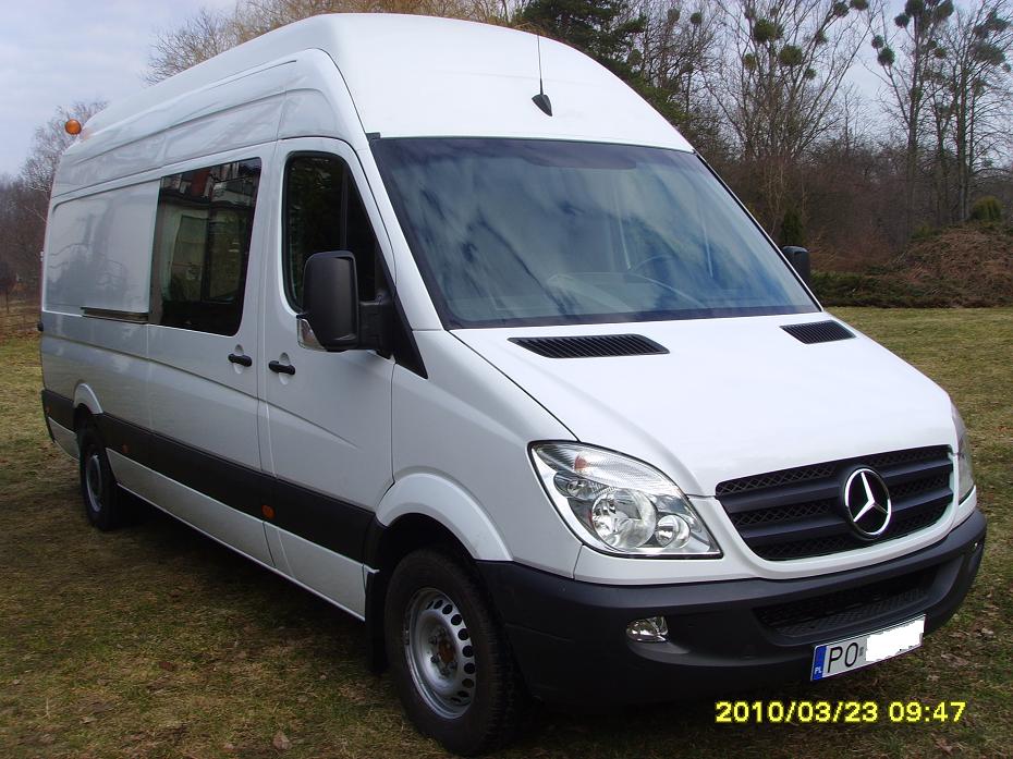 Biały, wysoki furgon Mercedes-Benz Sprinter na trawiastym terenie, widok z przodu i boku, z widoczną datą na dole zdjęcia.