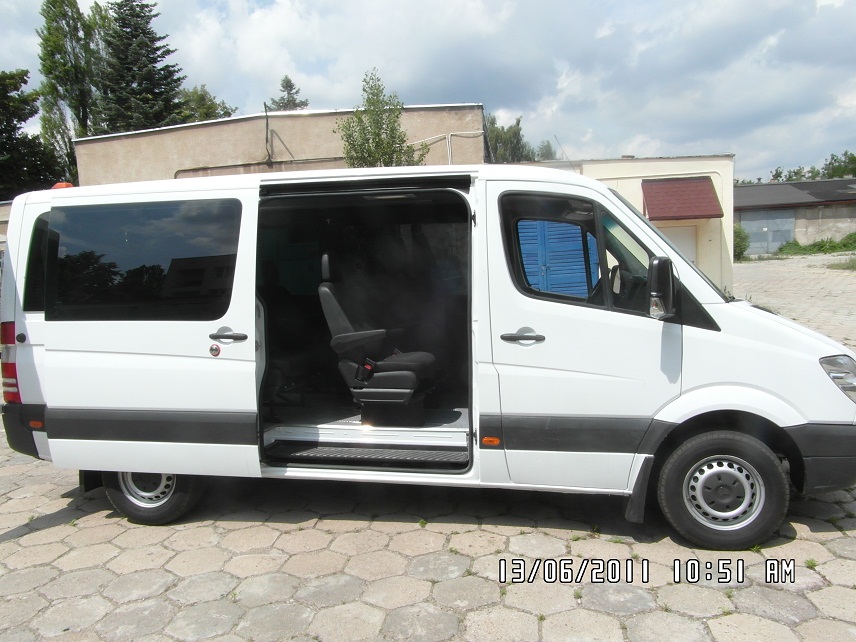 Biały bus z otwartymi przesuwnymi drzwiami, widoczne wnętrze z fotelem pasażera, zaparkowany na brukowanym placu.