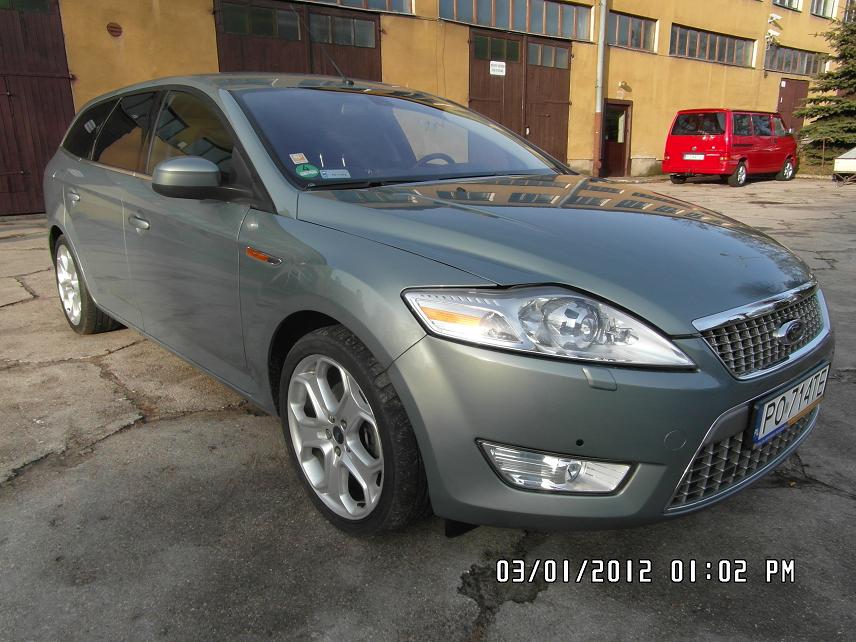 Szary samochód kombi marki Ford, zaparkowany przed budynkiem z żółtą elewacją i brązowymi bramami garażowymi, z czerwonym vanem w tle.