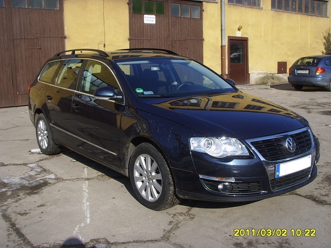 Czarny samochód kombi marki Volkswagen Passat zaparkowany na betonowym placu przed budynkiem z brązowymi drzwiami garażowymi; widoczne odbicie budynku w karoserii.