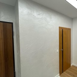 Rembudpro - Ściana z dekoracyjną, jasną strukturą przypominającą marmur, drewniane drzwi z czarnym paskiem, podłoga z jasnego drewna i marmurowe płytki podłogowe.