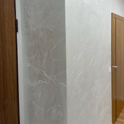 Rembudpro - Ściana pokryta dekoracyjną, jasną gładzią z subtelnym efektem marmuru, widoczna przy drewnianych drzwiach z ciemną ramą.