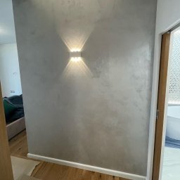 Rembudpro - Ściana z efektem dekoracyjnym w odcieniach szarości, oświetlona kinkietem z dwoma źródłami światła, widoczny fragment drewnianej podłogi i drzwi do łazienki z wzorzystą szybą.