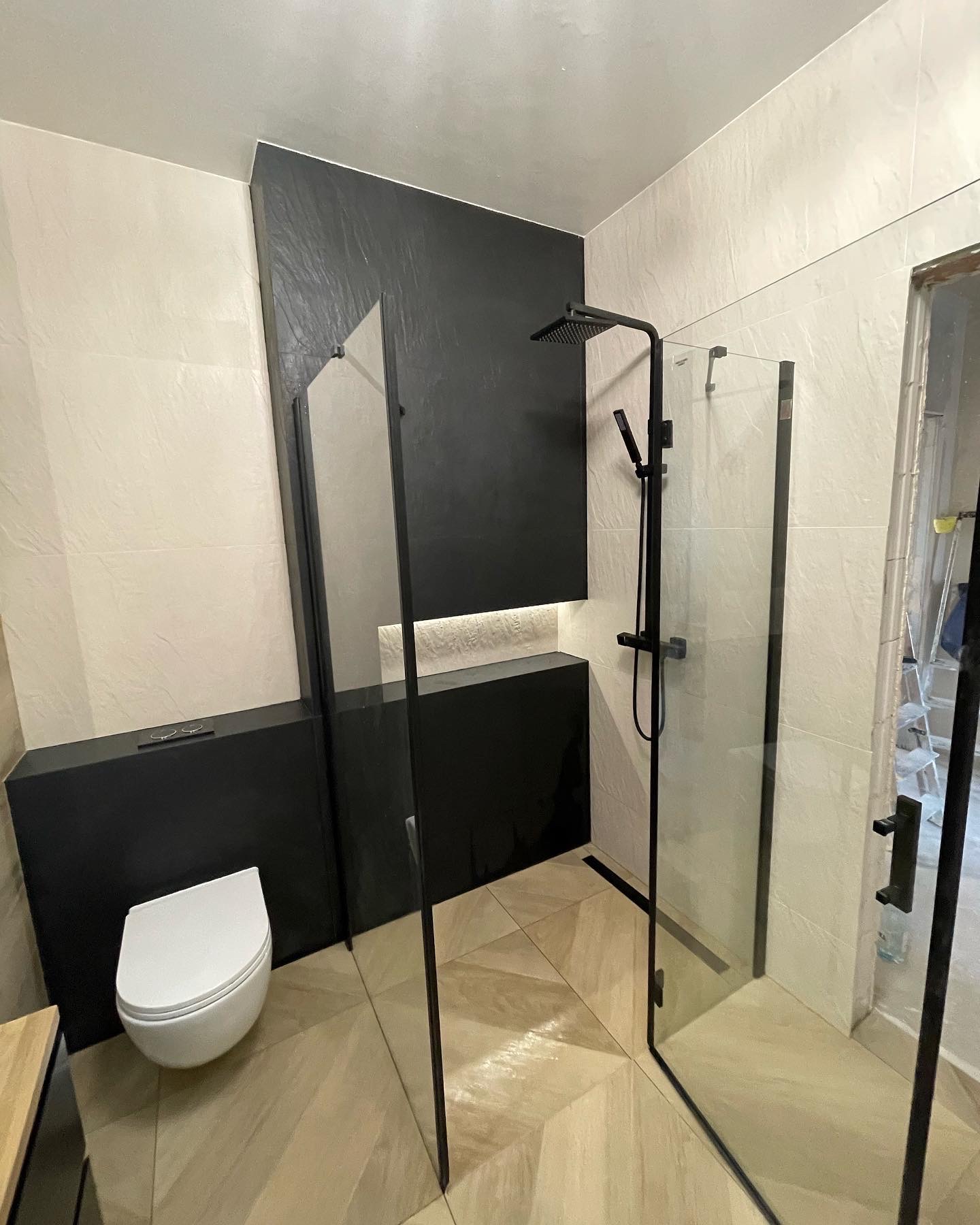 Nowoczesna łazienka z czarnymi akcentami: kabina prysznicowa typu walk-in z czarnymi profilami, deszczownica, podwieszana toaleta w zabudowie, beżowe płytki na ścianach i podłodze ułożone we wzór...