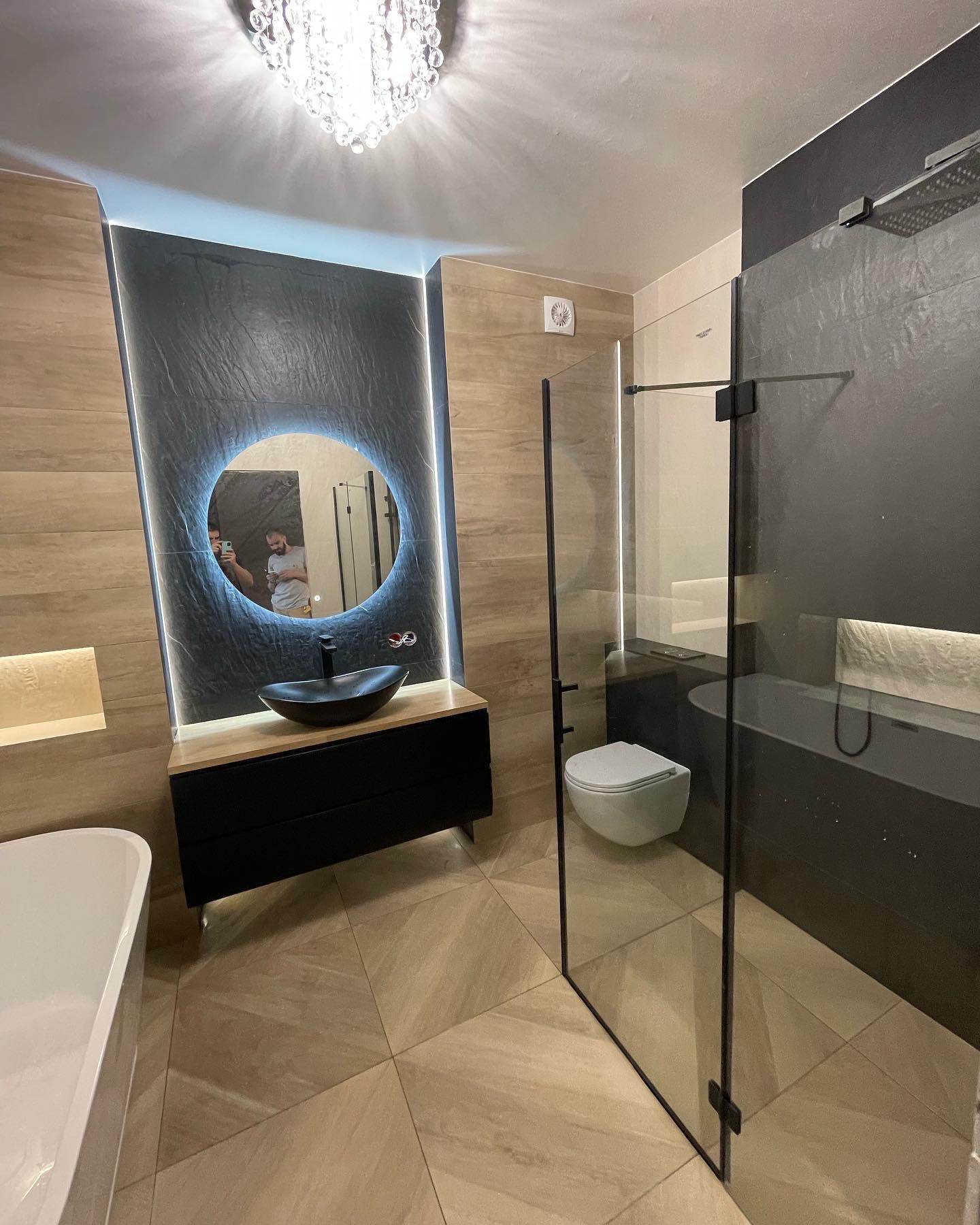 Nowoczesna łazienka z czarną umywalką w kształcie łzy, podświetlanym okrągłym lustrem i szklaną kabiną prysznicową walk-in z czarnymi elementami, oświetlona kryształowym żyrandolem.