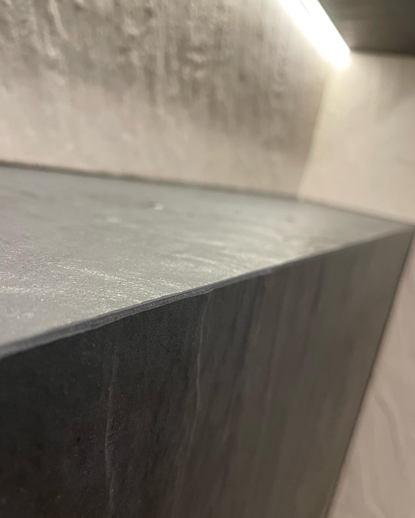 Szara półka z płytek w łazience, oświetlona od góry, z widoczną fakturą ściany w tle.