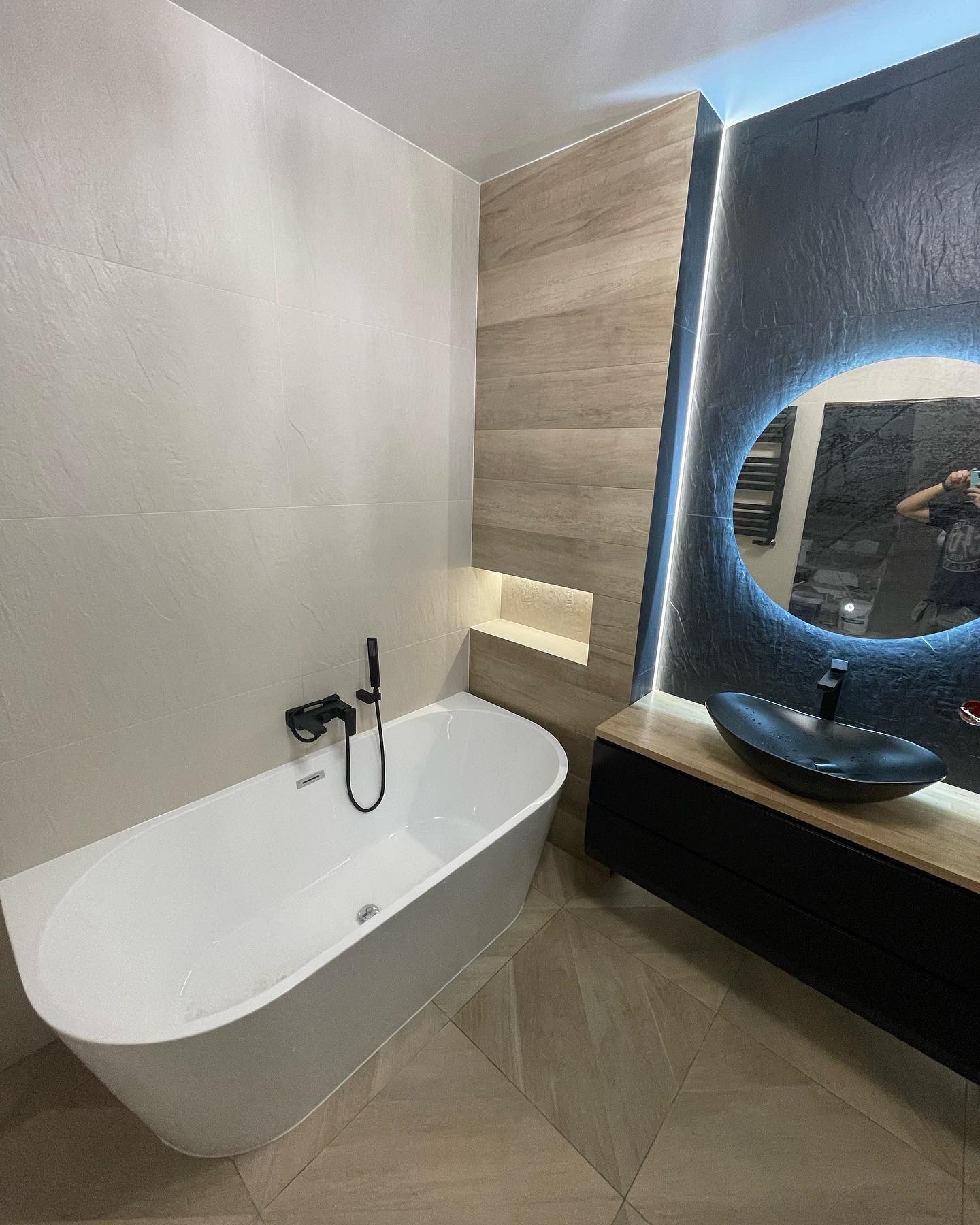 Nowoczesna łazienka z wolnostojącą białą wanną, czarną armaturą i umywalką w kształcie łzy na drewnianym blacie, oświetlone okrągłe lustro i szara ściana z efektem kamienia.