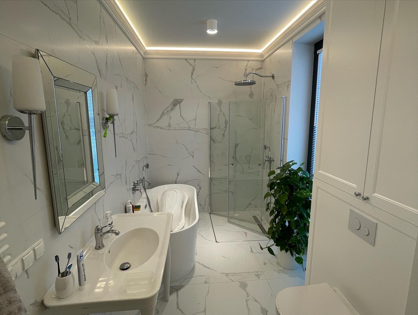 Elegancka łazienka w bieli i szarości z marmurowymi płytkami, podświetlanym sufitem, lustrem w srebrnej ramie, nowoczesną umywalką i zieloną rośliną doniczkową obok kabiny prysznicowej.