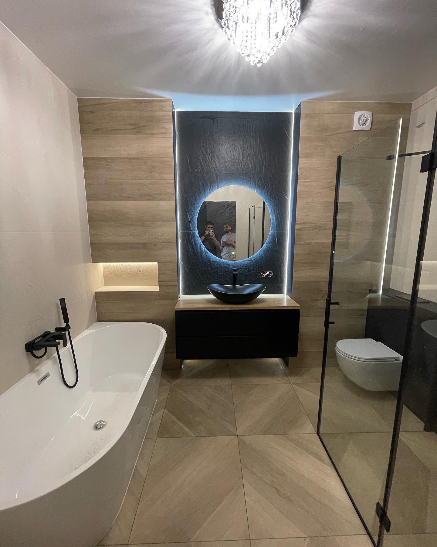 Nowoczesna łazienka z wolnostojącą wanną, czarną armaturą, okrągłym lustrem z podświetleniem LED i kabiną prysznicową ze szklanymi drzwiami.