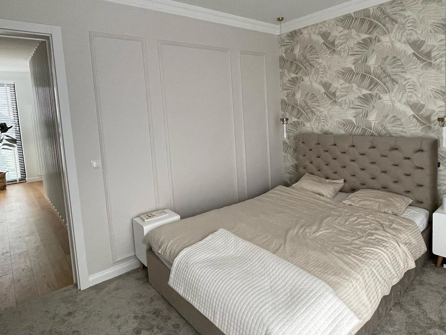 Beżowa sypialnia z tapetą w liście, pikowanym zagłówkiem i białymi panelami ściennymi, oświetlona lampami wiszącymi z przezroczystymi kloszami.