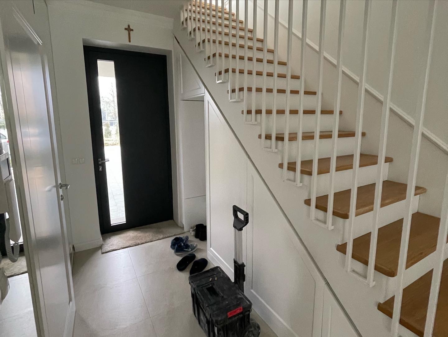 Jasne wnętrze domu w Warszawie z drewnianymi schodami na białych wspornikach, czarnymi drzwiami wejściowymi z krzyżem nad nimi i skrzynką narzędziową na podłodze.