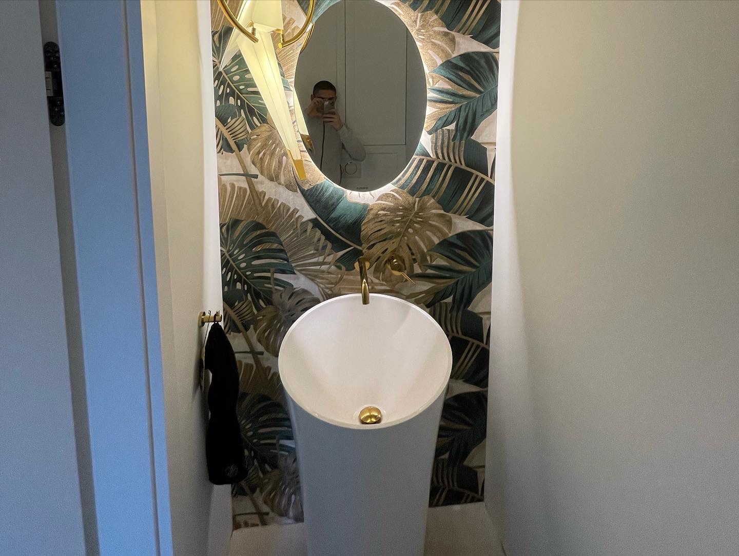 Nowoczesna łazienka z umywalką na postumencie, lustrem z podświetleniem LED i tapetą w motywy tropikalne, widoczne odbicie osoby robiącej zdjęcie.