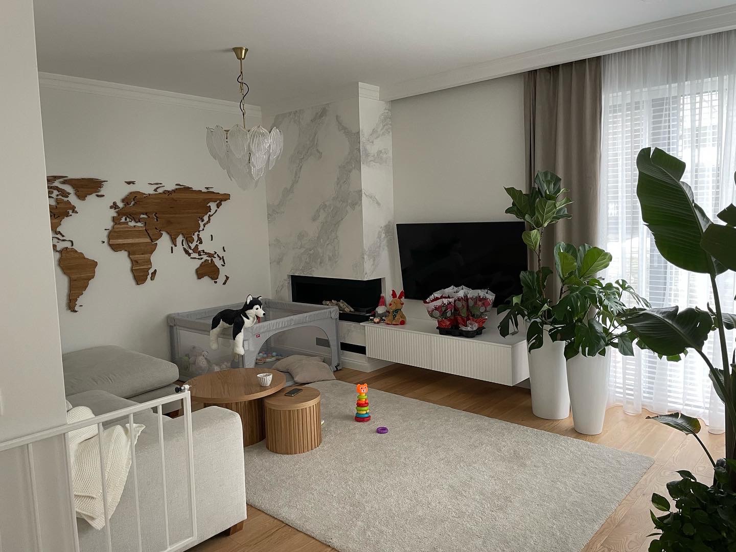 Jasne, przestronne wnętrze salonu z drewnianą mapą świata na ścianie, marmurowym kominkiem, białymi meblami, roślinami w donicach i dziecięcym kojcem z pluszowym psem.