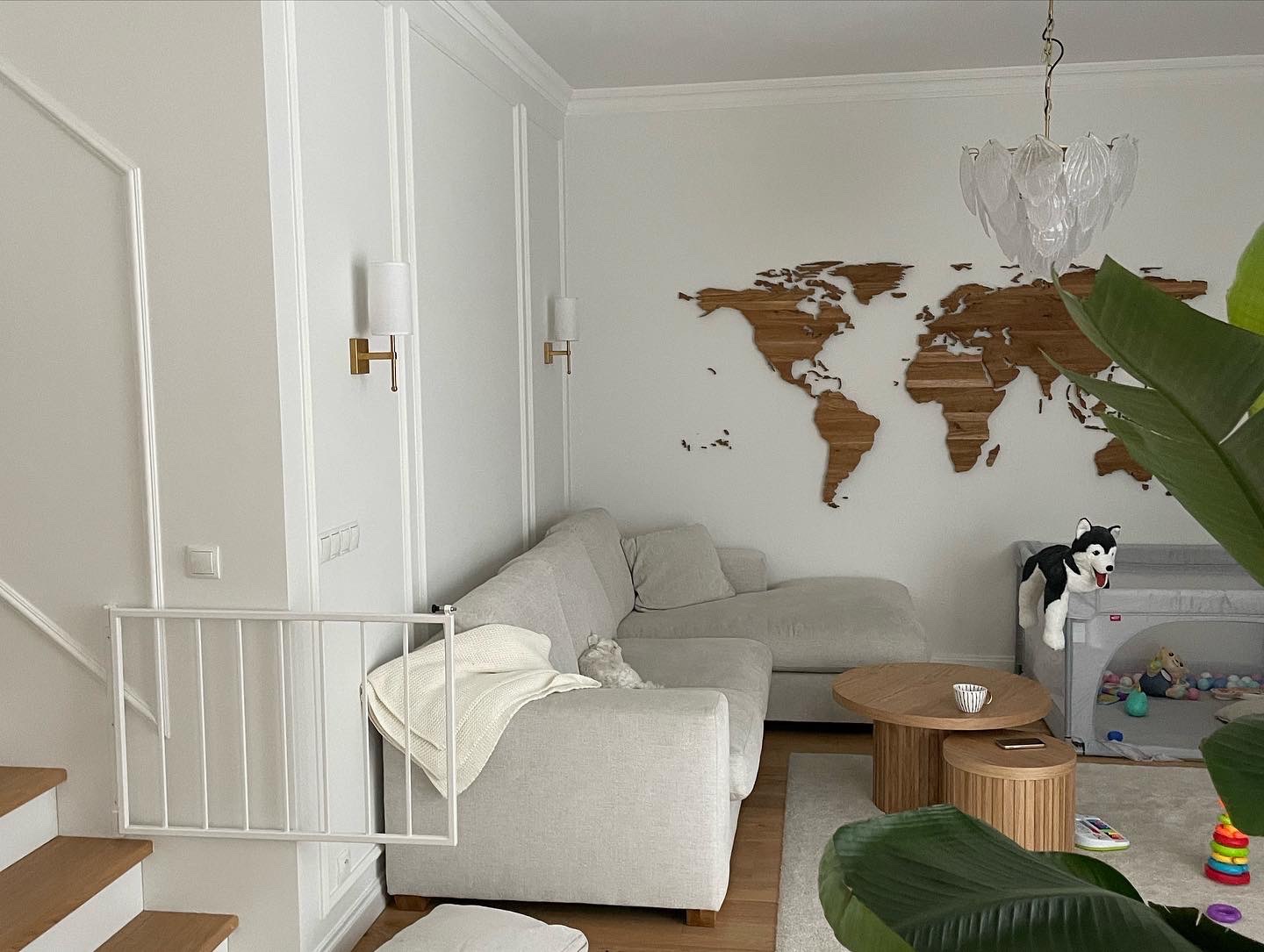 Jasne wnętrze salonu z narożną sofą, drewnianą mapą świata na ścianie i dekoracyjnymi listwami ściennymi, oświetlone złotymi kinkietami.