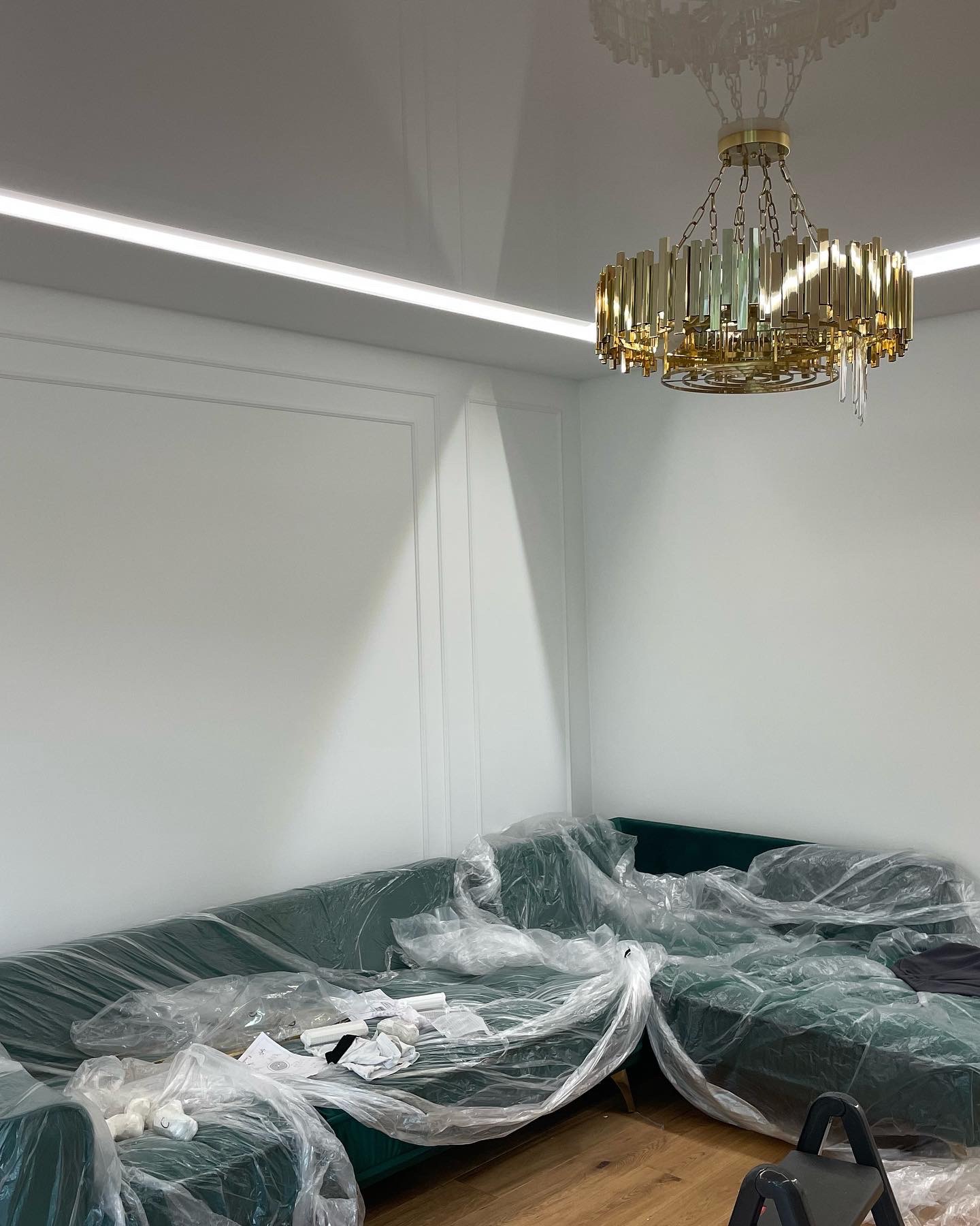 Pokój w trakcie prac wykończeniowych: zielona sofa zabezpieczona folią, złoty żyrandol w stylu art deco, białe ściany z dekoracyjnymi listwami i oświetleniem LED.