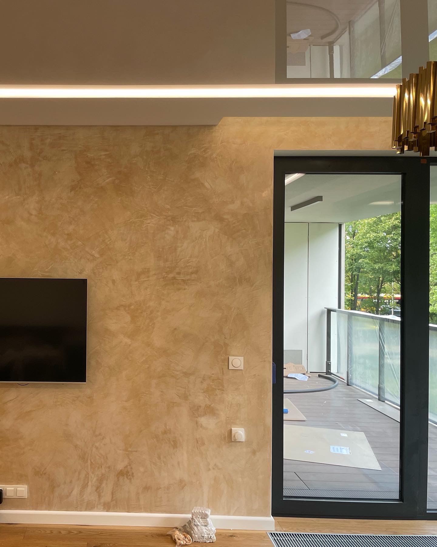 Ściana z efektem dekoracyjnym w odcieniach beżu i brązu, oświetlenie LED podsufitowe, czarny telewizor na ścianie, wyjście na balkon z widokiem na zieleń, fragment złotej lampy wiszącej.