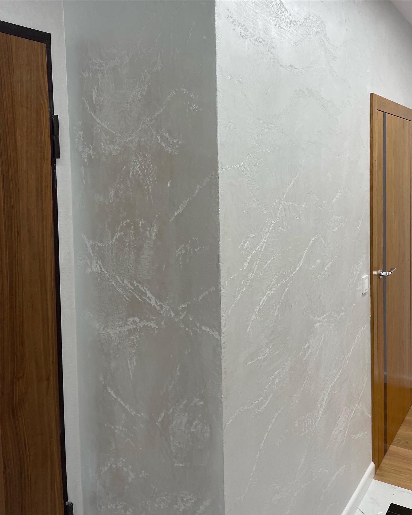 Ściana pokryta dekoracyjną, jasną gładzią z subtelnym efektem marmuru, widoczna przy drewnianych drzwiach z ciemną ramą.