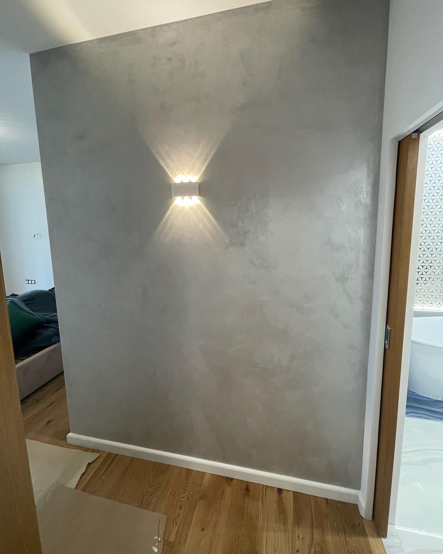 Ściana z efektem dekoracyjnym w odcieniach szarości, oświetlona kinkietem z dwoma źródłami światła, widoczny fragment drewnianej podłogi i drzwi do łazienki z wzorzystą szybą.