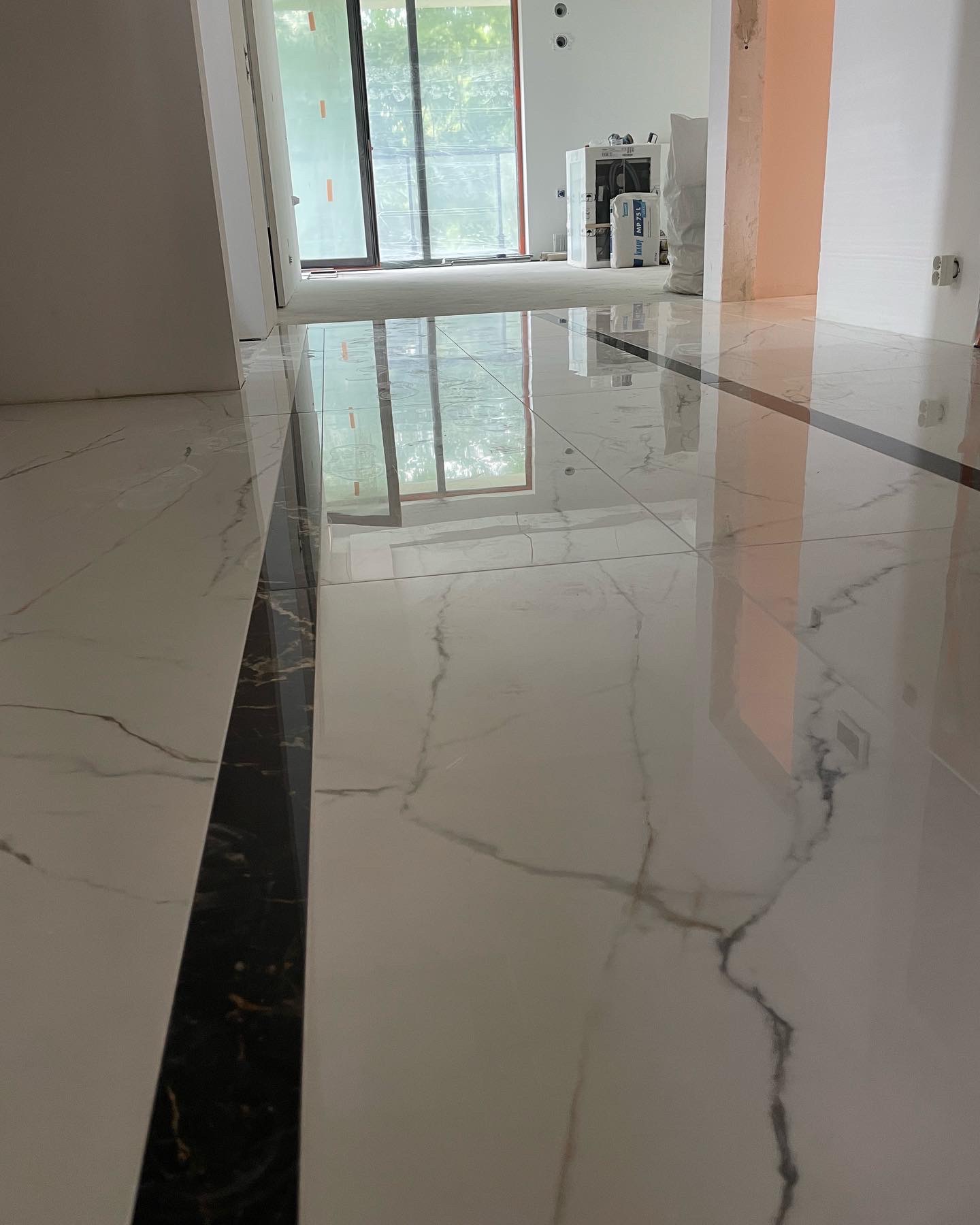 Błyszcząca, biała marmurowa podłoga z czarnym pasem dekoracyjnym, widok na przesuwne drzwi balkonowe i materiały budowlane w tle, wnętrze w trakcie wykańczania.