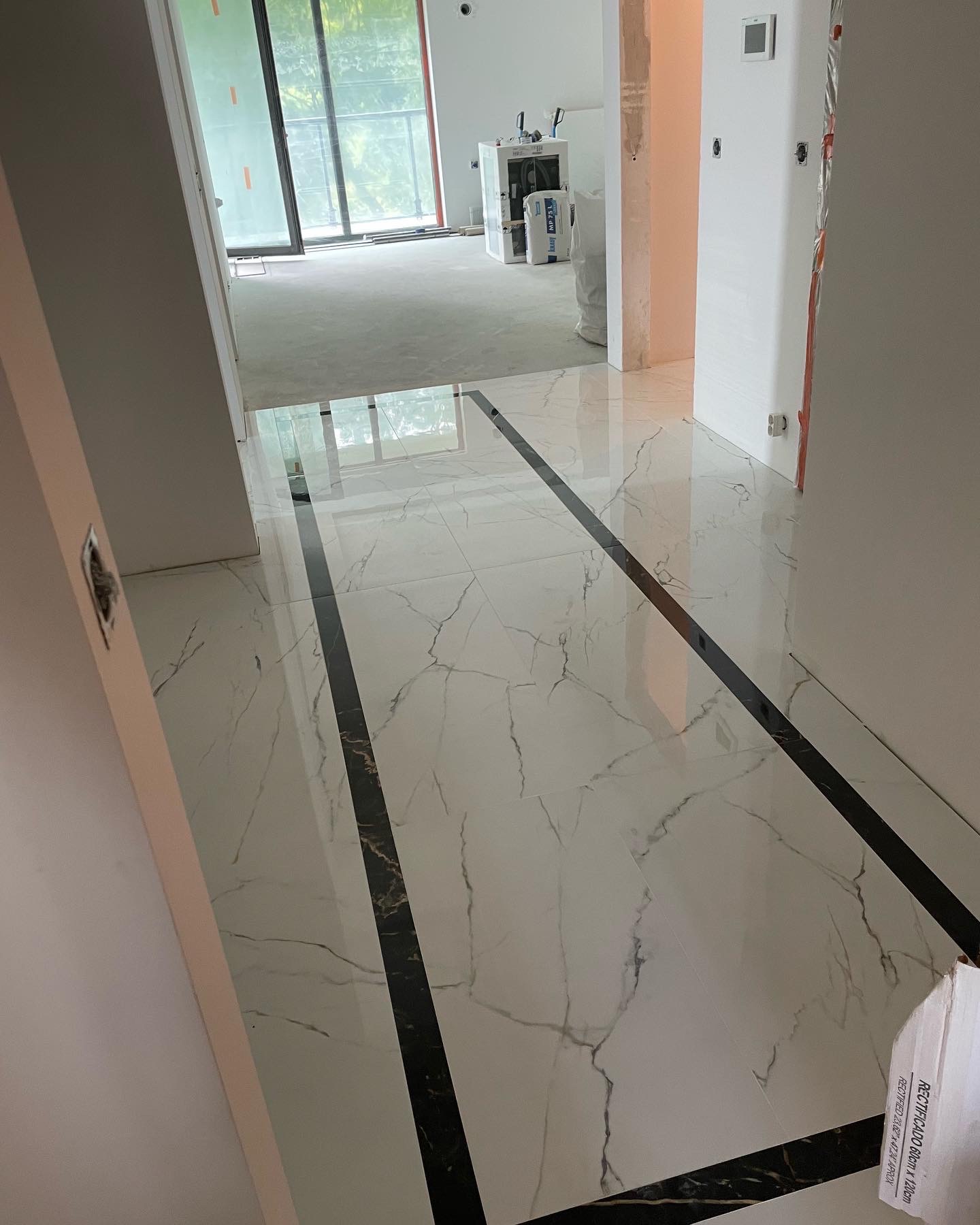 Błyszcząca podłoga z białych płytek imitujących marmur z czarnymi pasami dekoracyjnymi, widok na otwartą przestrzeń z workami materiałów budowlanych w tle.