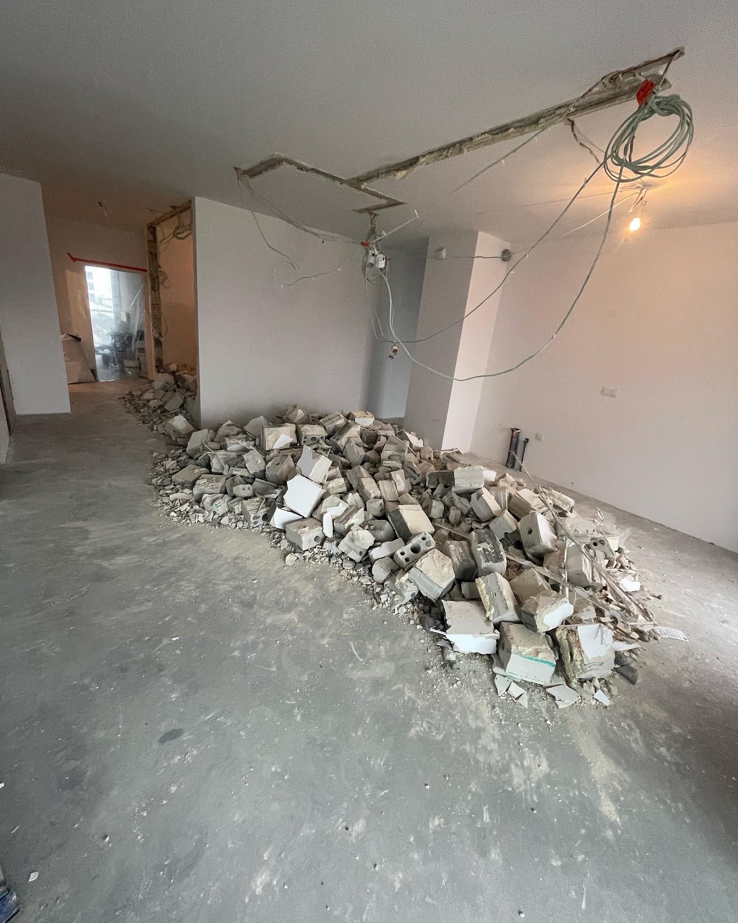 Stos gruzu i resztek materiałów budowlanych na szarym betonowym podłożu w trakcie prac rozbiórkowych wewnątrz pomieszczenia, z widocznymi instalacjami elektrycznymi zwisającymi z sufitu i otwartym...