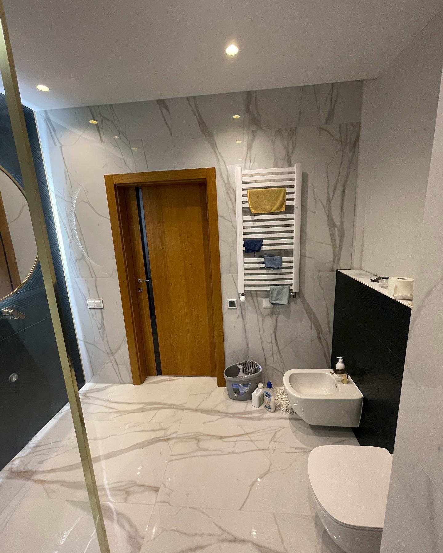 Nowoczesna łazienka z marmurowymi płytkami, drewnianymi drzwiami, białym grzejnikiem drabinkowym z ręcznikami, bidetem i wiszącą toaletą.