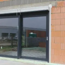 Montaż okna przesuwnego z szarą ramą i zewnętrzną roletą, widoczne puste przestrzenie w murze z cegły krat&oacute;wki oraz elementy instalacji ogrzewania podłogowego wewnątrz budynku.