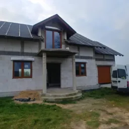 Dom w stanie surowym zamkniętym w Ostrowie Wielkopolskim z zamontowanymi oknami z brązowymi ramami i częściowo zamontowanymi roletami zewnętrznymi, widoczna brama garażowa i dwa samochody dostawcze.