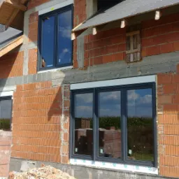 Budynek w trakcie budowy z zamontowanymi oknami w grafitowych ramach, widoczne bloczki ceramiczne i fragmenty drewnianej konstrukcji dachu.