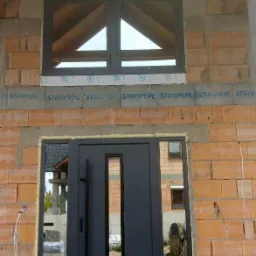 Montaż ciemnoszarych drzwi zewnętrznych z wąskim przeszkleniem i okna z trójkątnym szprosem w surowym stanie budynku z cegły.