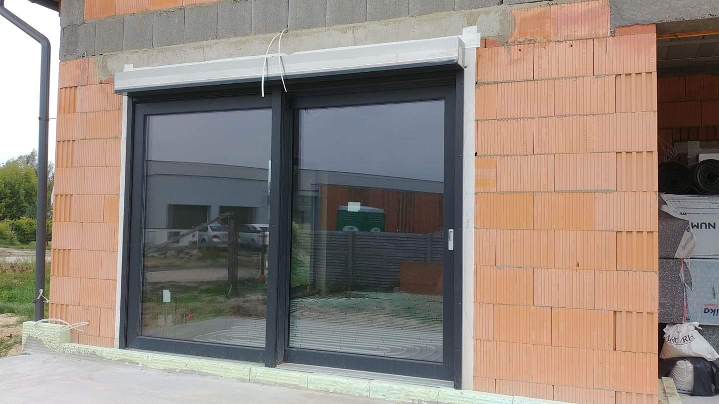 Montaż okna przesuwnego z szarą ramą i zewnętrzną roletą, widoczne puste przestrzenie w murze z cegły kratówki oraz elementy instalacji ogrzewania podłogowego wewnątrz budynku.