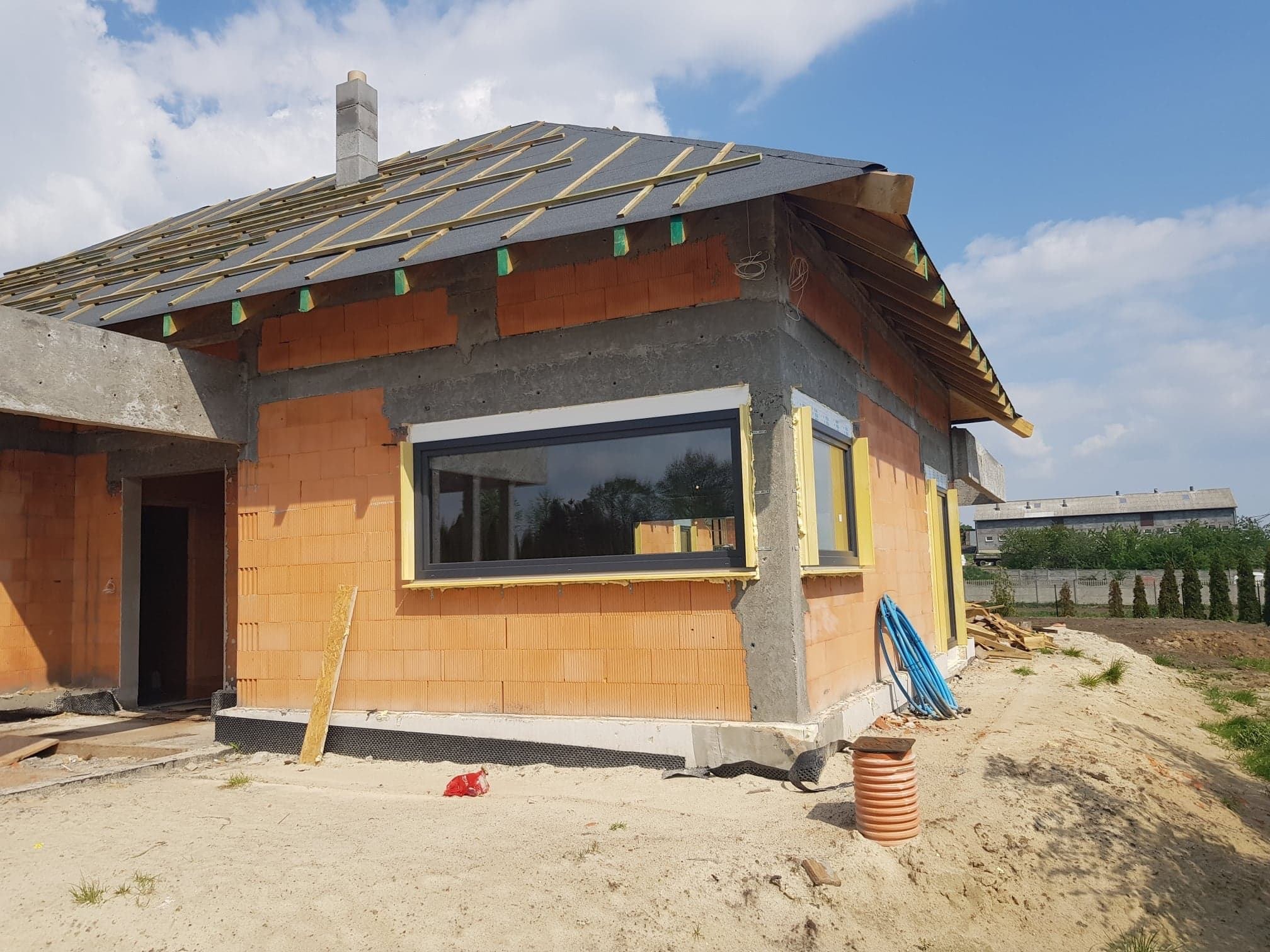 Surowy stan domu jednorodzinnego w trakcie budowy: widoczne okna PCV z czarnymi ramami, murowane ściany z pustaków ceramicznych, przygotowane podłoże pod dach z papą, teren wokół budynku w stanie...