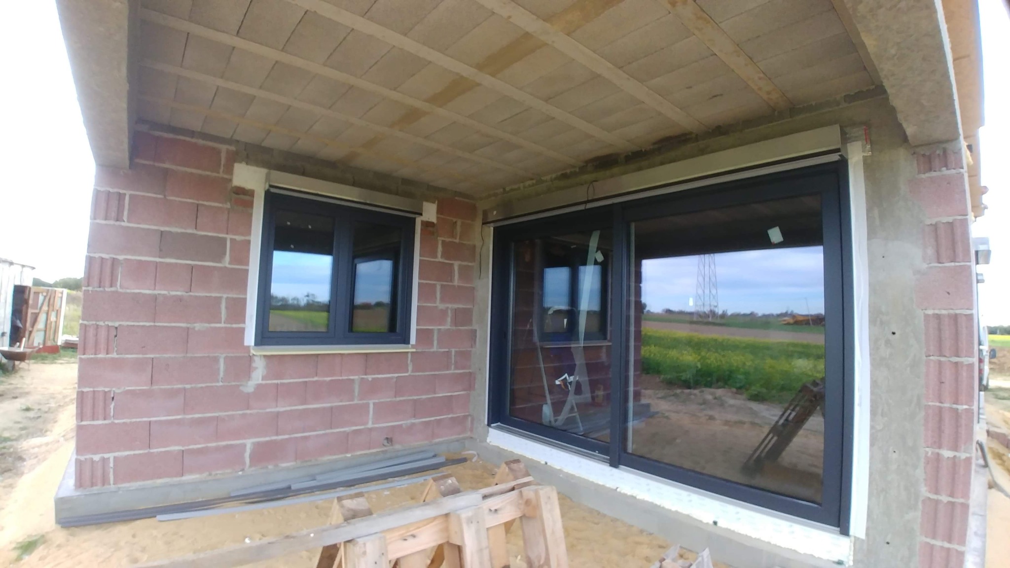 Surowy stan budynku w trakcie budowy z zamontowanymi oknami w ciemnych ramach, widok z zewnątrz, paleta drewniana na piasku przed oknami.